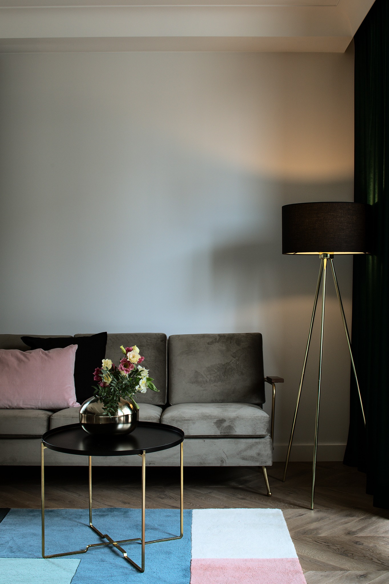 Stylowy salon: szara sofa z poduszkami, stolik kawowy z kwiatami, lampa trójnóg i kolorowy dywan na parkiecie w jodełkę. Eleganckie wnętrze.