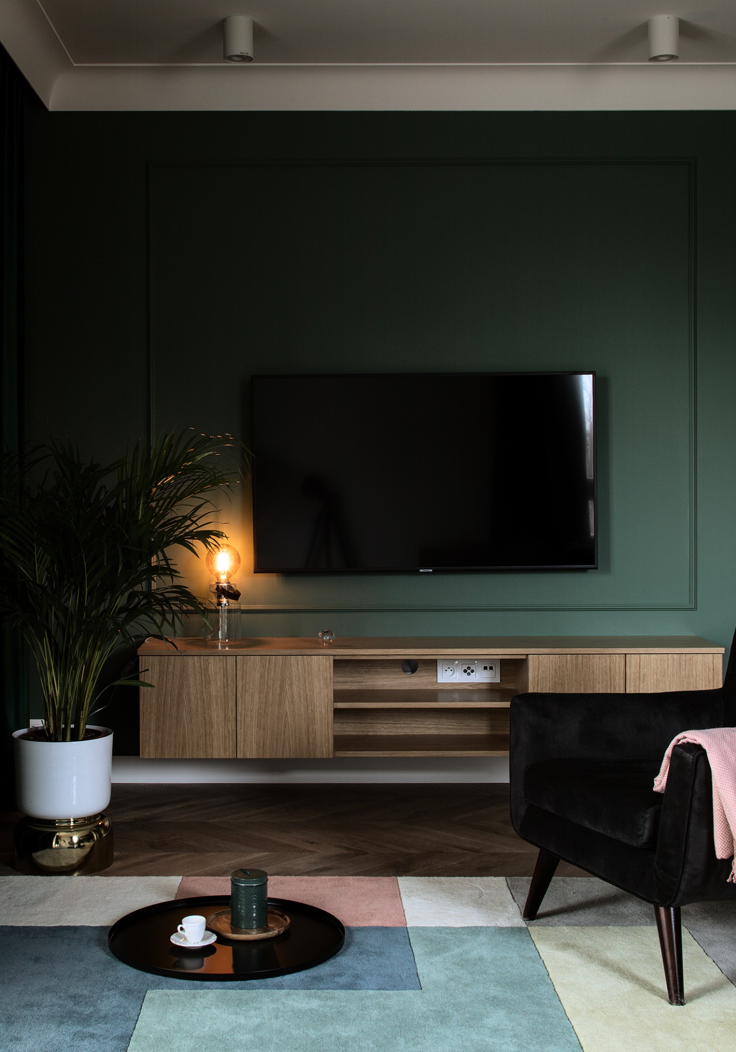 Stylowy salon z zieloną ścianą, drewnianą szafką RTV, czarnym fotelem i kolorowym dywanem. Lampa z żarówką Edisona dodaje ciepłego światła.