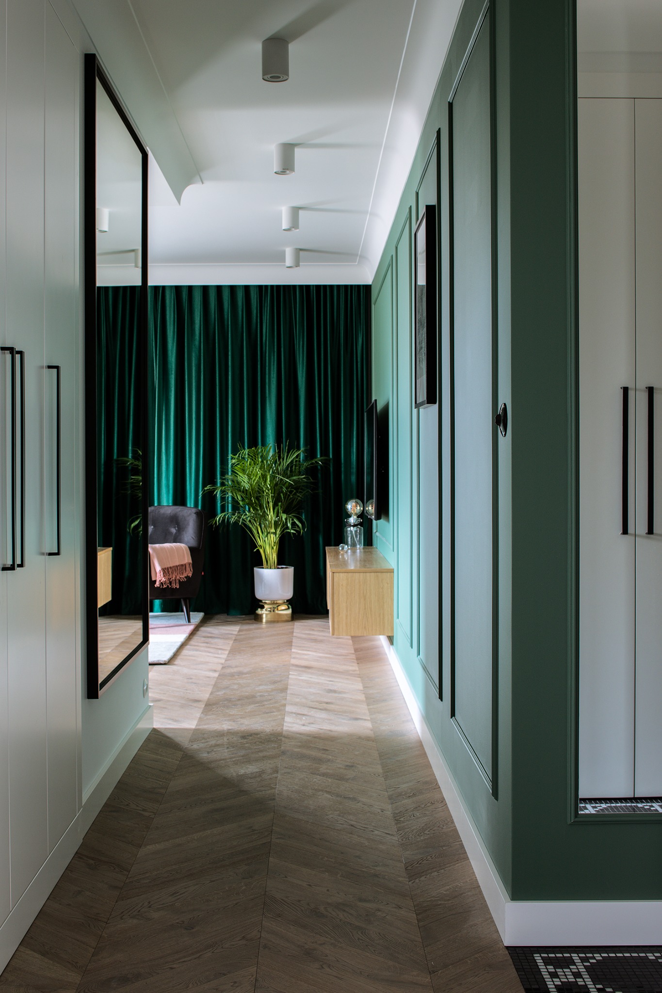 Stylowy korytarz z zielonymi ścianami, drewnianą podłogą w jodełkę i rośliną w złotej donicy. W tle zasłony w kolorze butelkowej zieleni i designerskie oświetlenie.