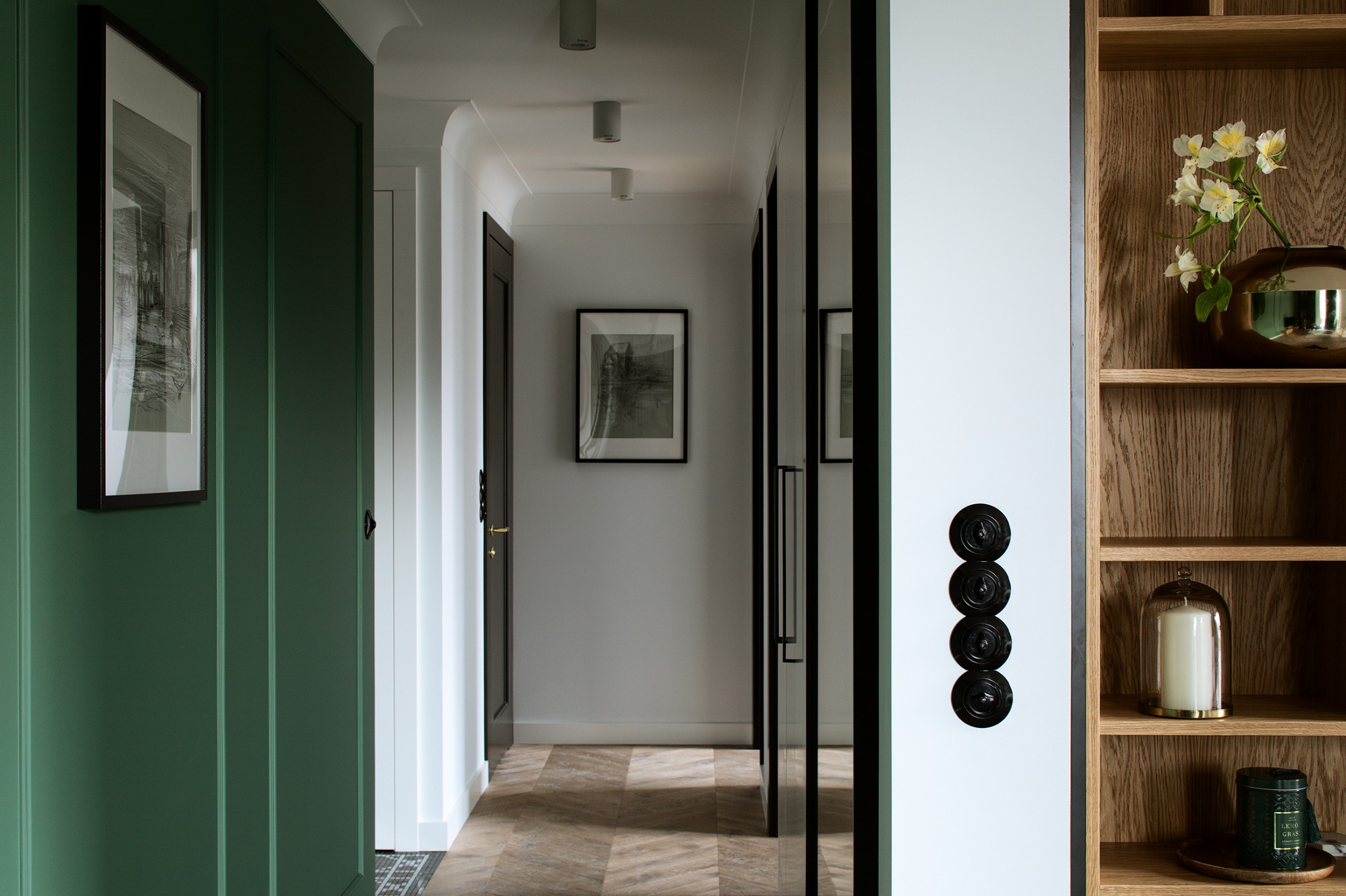 Stylowy korytarz z zieloną boazerią, lustrem w czarnej ramie i drewnianym regałem z dekoracjami. Parkiet w jodełkę i czarne włączniki dodają elegancji.