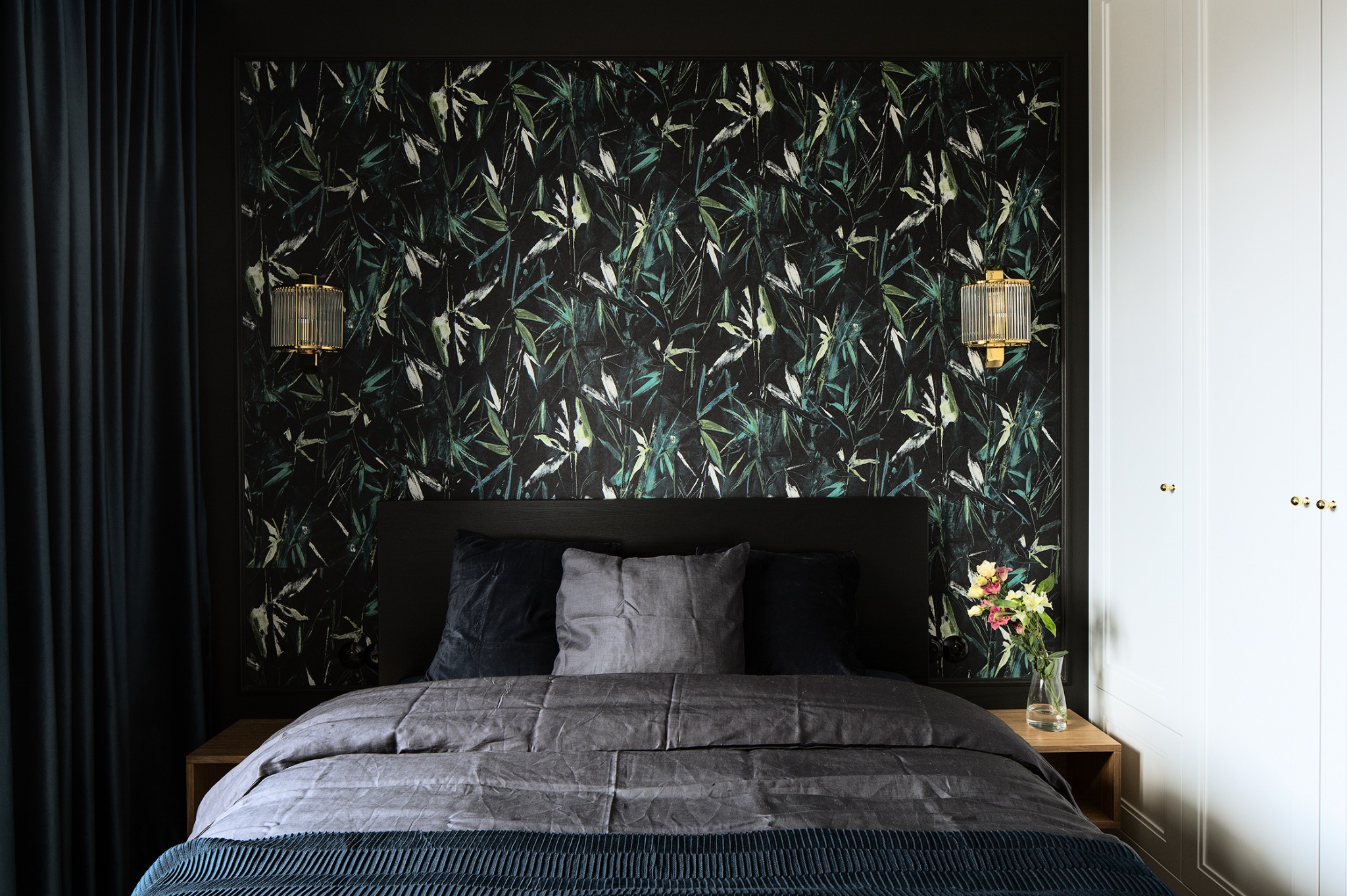 Sypialnia z tapetą w motywy roślinne, oświetlona złotymi kinkietami. Na łóżku poduszki w odcieniach szarości i granatowa narzuta. Obok biała szafa i kwiaty w wazonie.