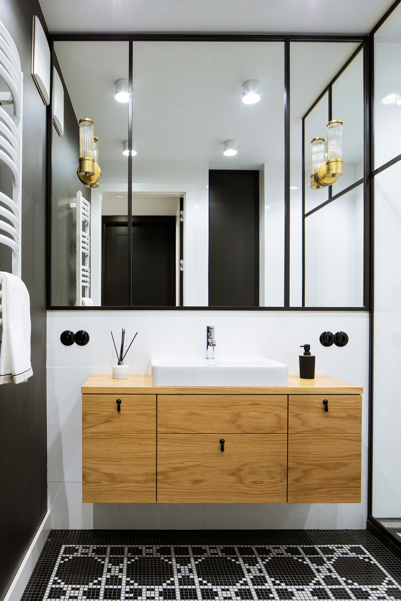 Nowoczesna łazienka z drewnianą szafką pod umywalką, czarno-białą mozaiką na podłodze i dużym lustrem w czarnej ramie. Minimalistyczny design.