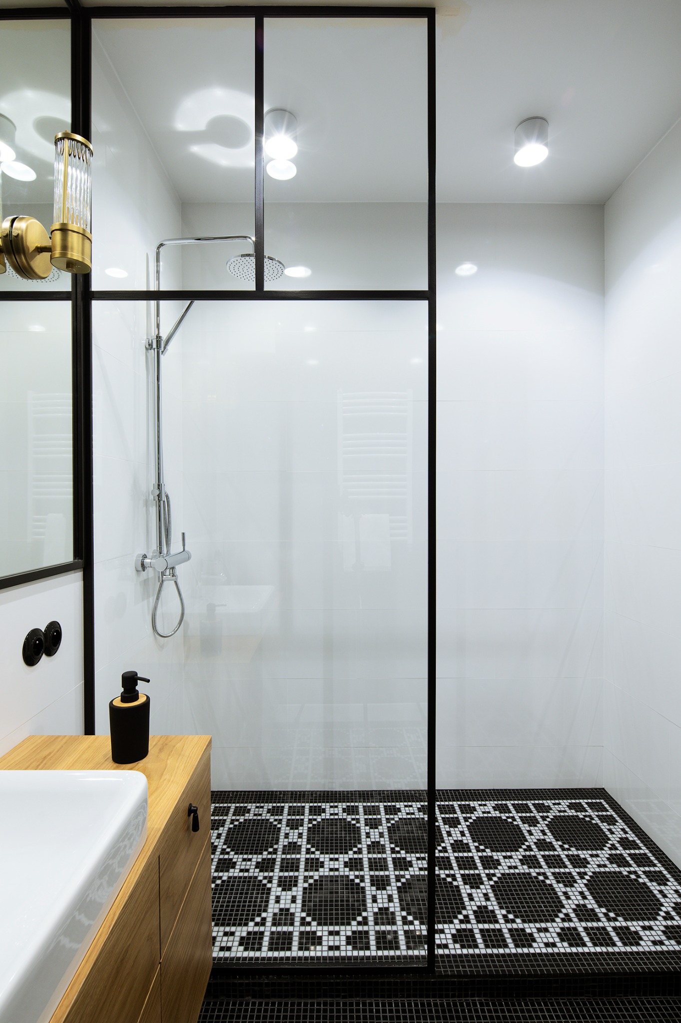 Nowoczesna łazienka z kabiną typu walk-in, czarno-białą mozaiką podłogową i drewnianą szafką pod umywalką. Minimalistyczny design z akcentami złota.