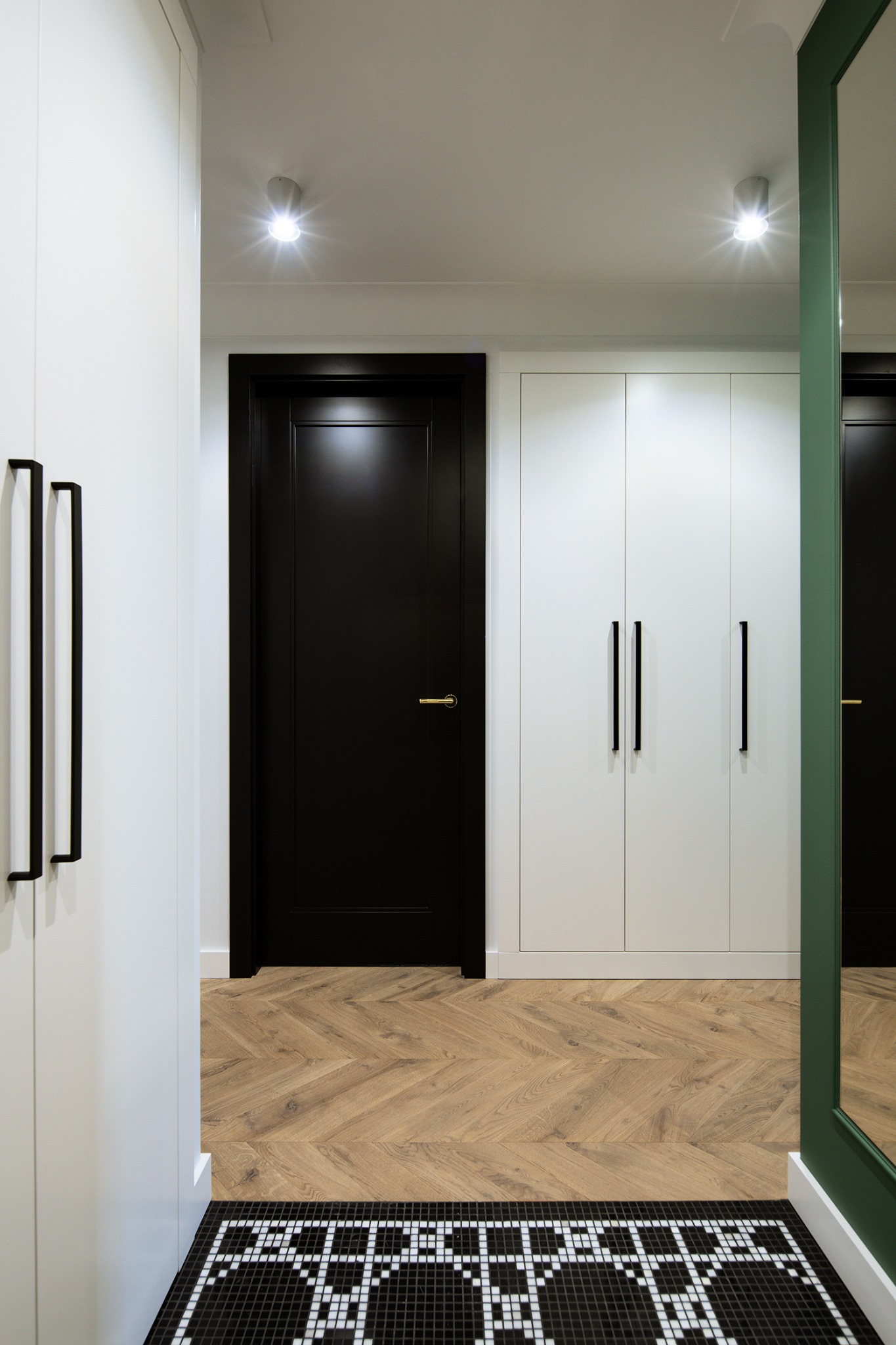 Nowoczesny przedpokój: czarne drzwi, biała szafa z podłużnymi uchwytami, parkiet w jodełkę i czarno-biała mozaika przy wejściu. Minimalistyczny design.