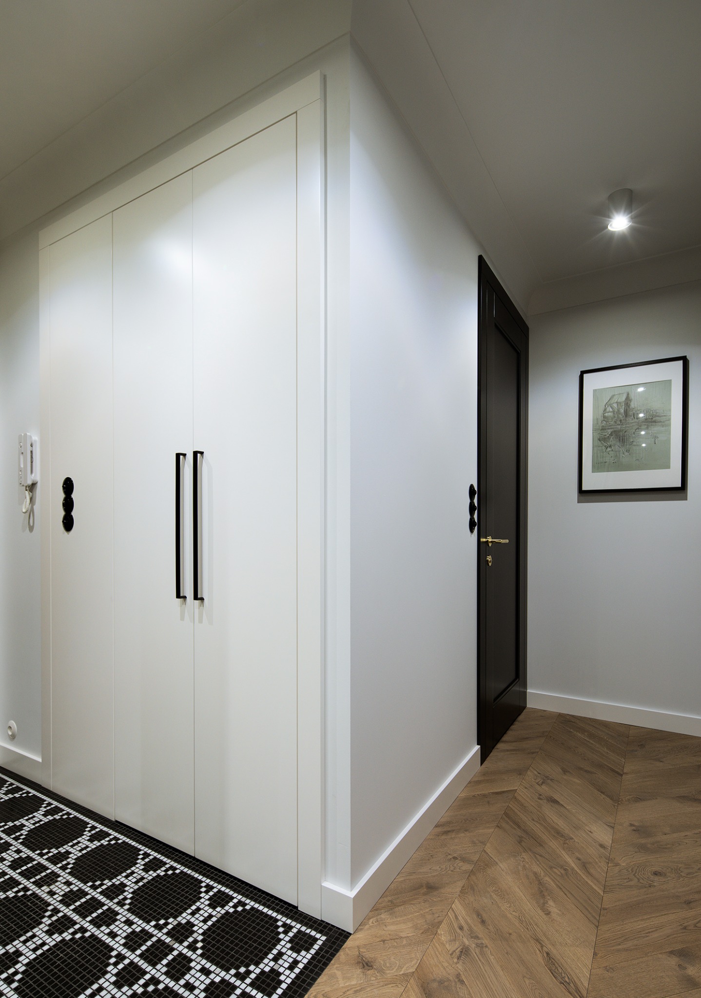 Nowoczesny przedpokój: biała szafa w zabudowie z czarnymi uchwytami, mozaika na podłodze i parkiet w jodełkę, czarne drzwi i obraz na ścianie.