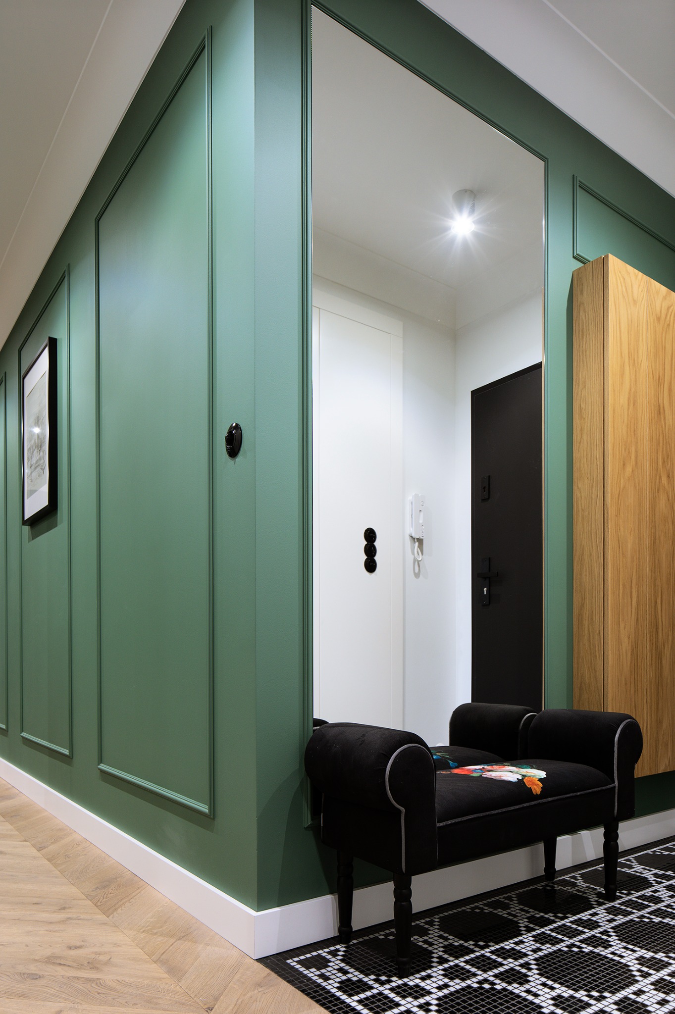 Stylowy przedpokój z zielonymi panelami ściennymi, czarną ławeczką z motywem kwiatowym i geometryczną posadzką. Widoczne drzwi wejściowe i szafa w zabudowie.