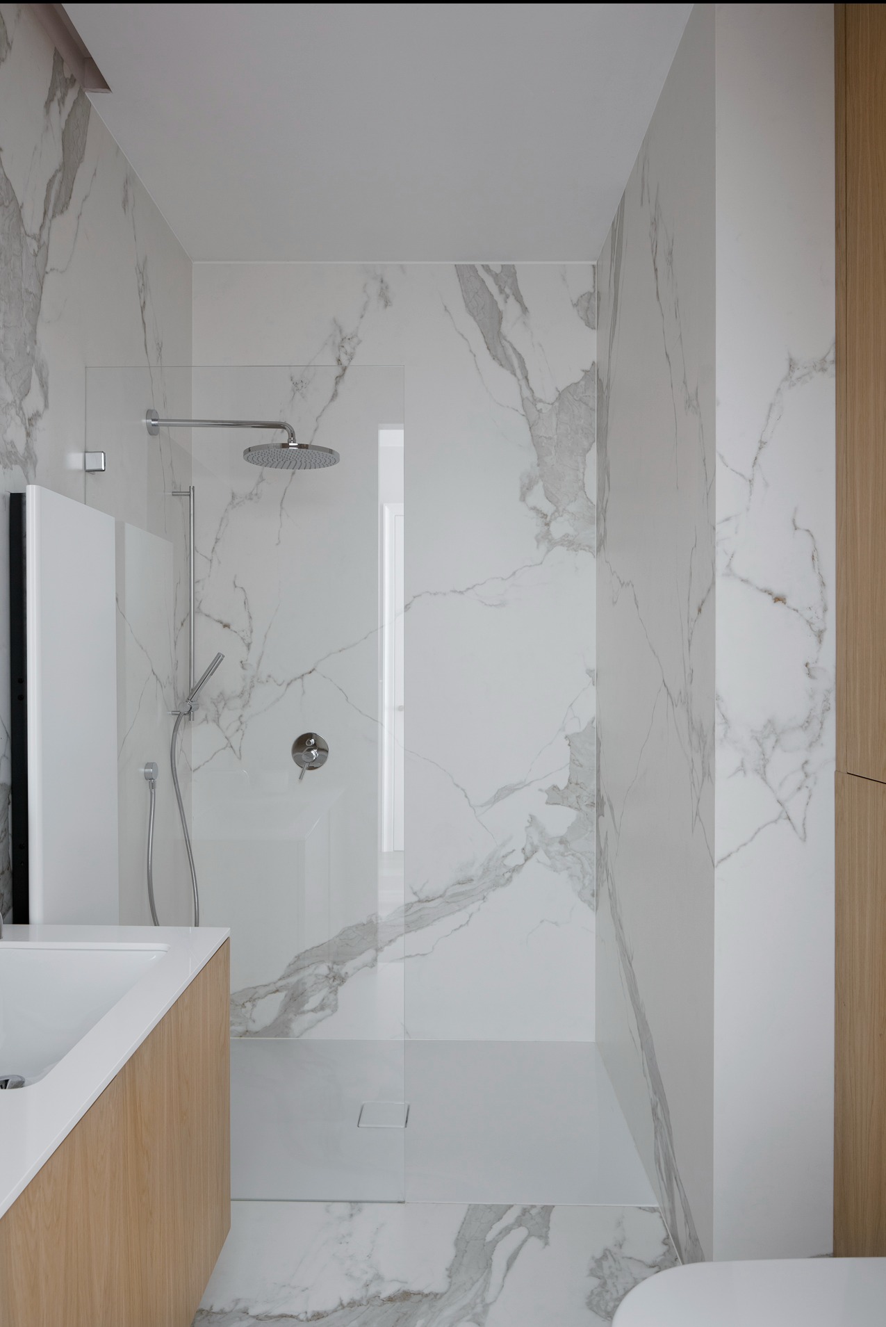 Nowoczesna łazienka z kabiną walk-in, wykończona białym marmurem. Widoczna armatura chromowana, umywalka z drewnianą szafką i odpływ liniowy w podłodze.