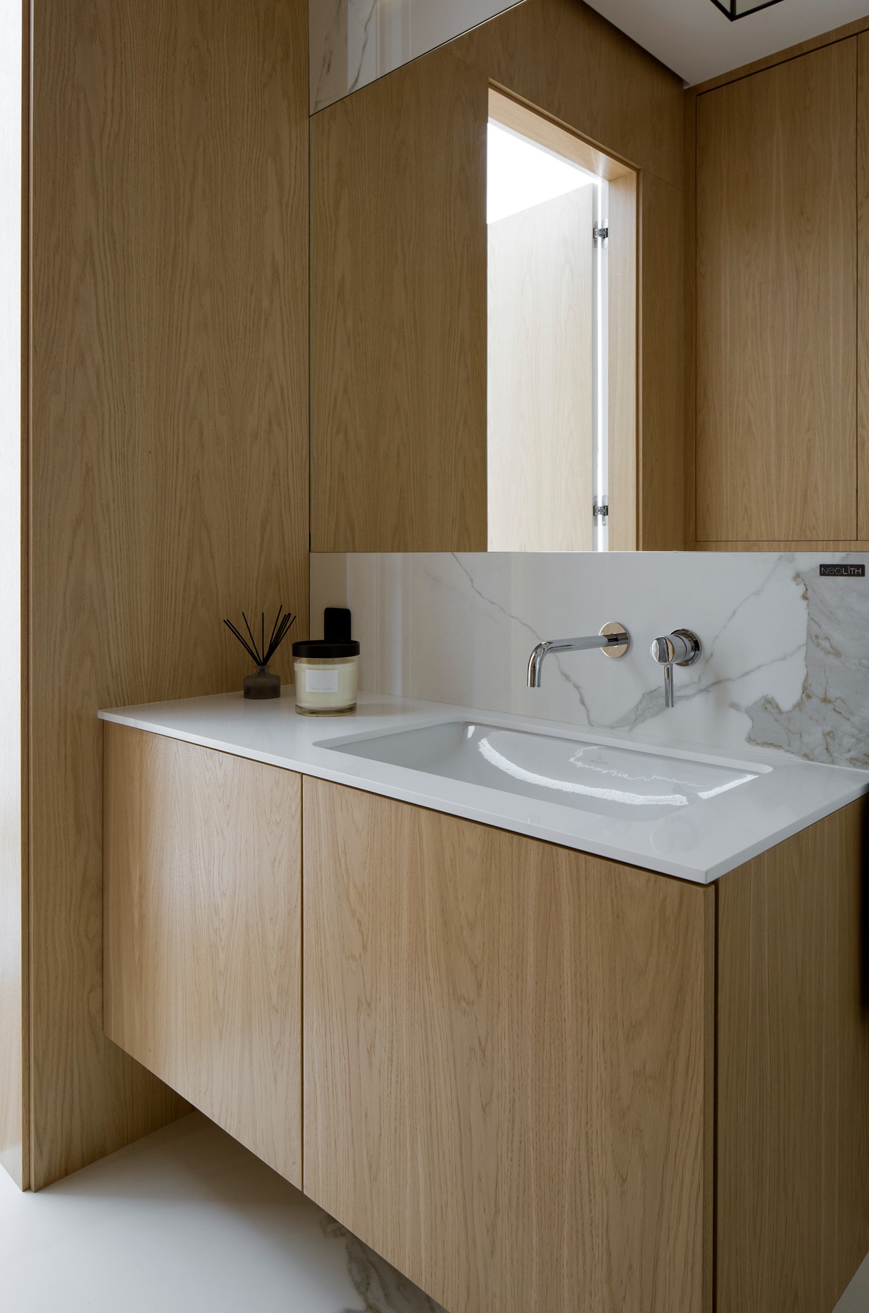 Minimalistyczna łazienka: drewniana szafka pod umywalką z białym blatem i marmurowym panelem. Chromowana bateria, świeca i zapach w tle.