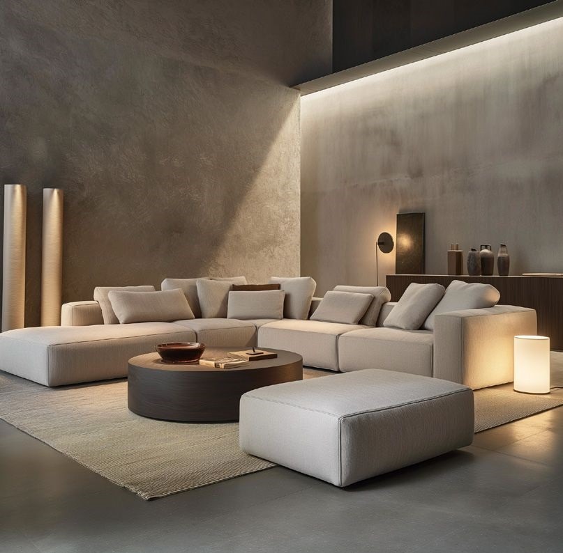 Elegancki salon z modułową sofą w odcieniach beżu, okrągłym stolikiem kawowym i minimalistycznym oświetleniem. Ściany w surowym stylu tworzą nowoczesny klimat.