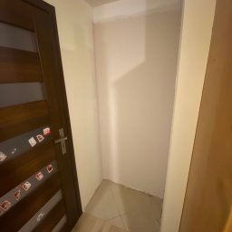 Dariusz Dębek - Wnęka w przedpokoju po malowaniu, widoczna ściana w odcieniach beżu, brązowe drzwi z naklejkami i fragment podłogi z jasnych płytek. Świeżo odmalowane ściany.