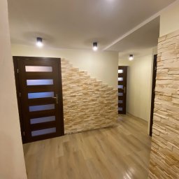 Dariusz Dębek - Korytarz z drewnianą podłogą i dekoracyjną ścianą z kamienia. Widoczne drzwi z przeszkleniami i oświetlenie sufitowe. Nietypowe wykończenie ściany z kamienia.