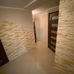 Dariusz Dębek - Korytarz z dekoracyjną ścianą z kamienia i drewnianymi drzwiami z przeszkleniami. Beżowe płytki podłogowe. Nowoczesne wykończenie wnętrza.