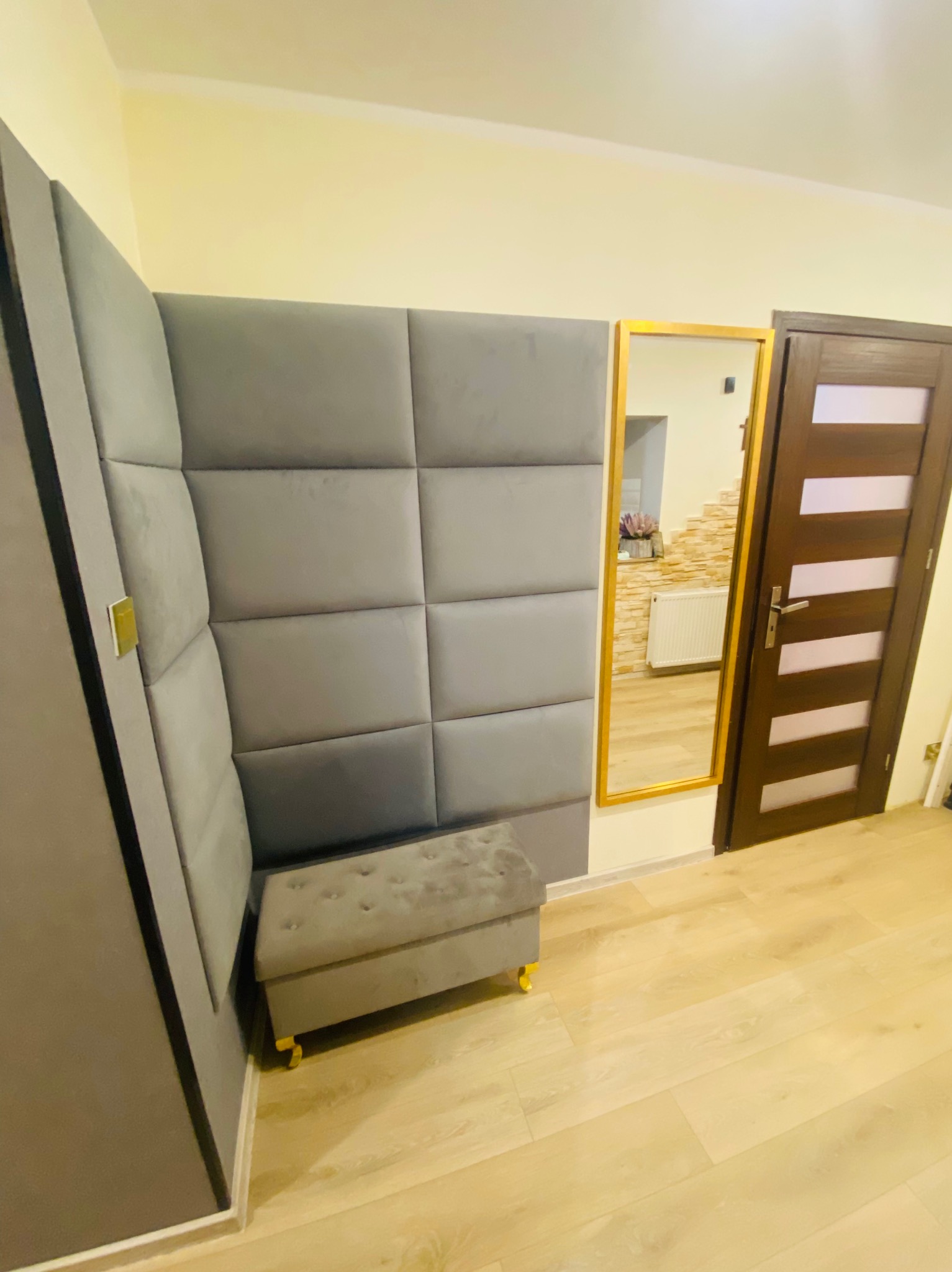 Szara, tapicerowana ściana z panelami w kwadraty, pufa z guzikami i lustro w złotej ramie, tworzące elegancki wystrój przedpokoju z drewnianą podłogą i drzwiami.