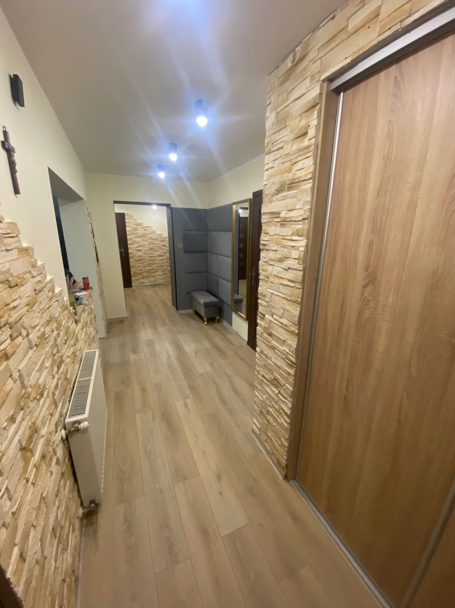 Wykończony korytarz z kamienną okładziną ściany, drewnianą podłogą i przesuwnymi drzwiami. Widoczny fragment grzejnika i dekoracyjny panel ścienny z siedziskiem w tle.