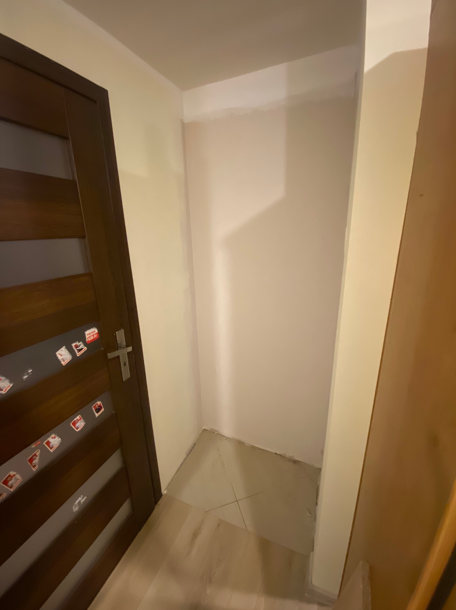 Wnęka w przedpokoju po malowaniu, widoczna ściana w odcieniach beżu, brązowe drzwi z naklejkami i fragment podłogi z jasnych płytek. Świeżo odmalowane ściany.