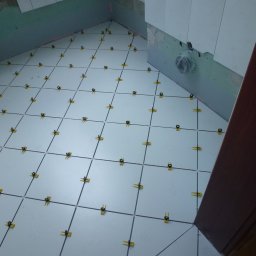 RAFAŁ GAJEWSKI - Białe płytki podłogowe w łazience, ułożone z użyciem żółtych krzyżyków dystansowych. Widoczne przygotowane ściany i fragment futryny drzwi.