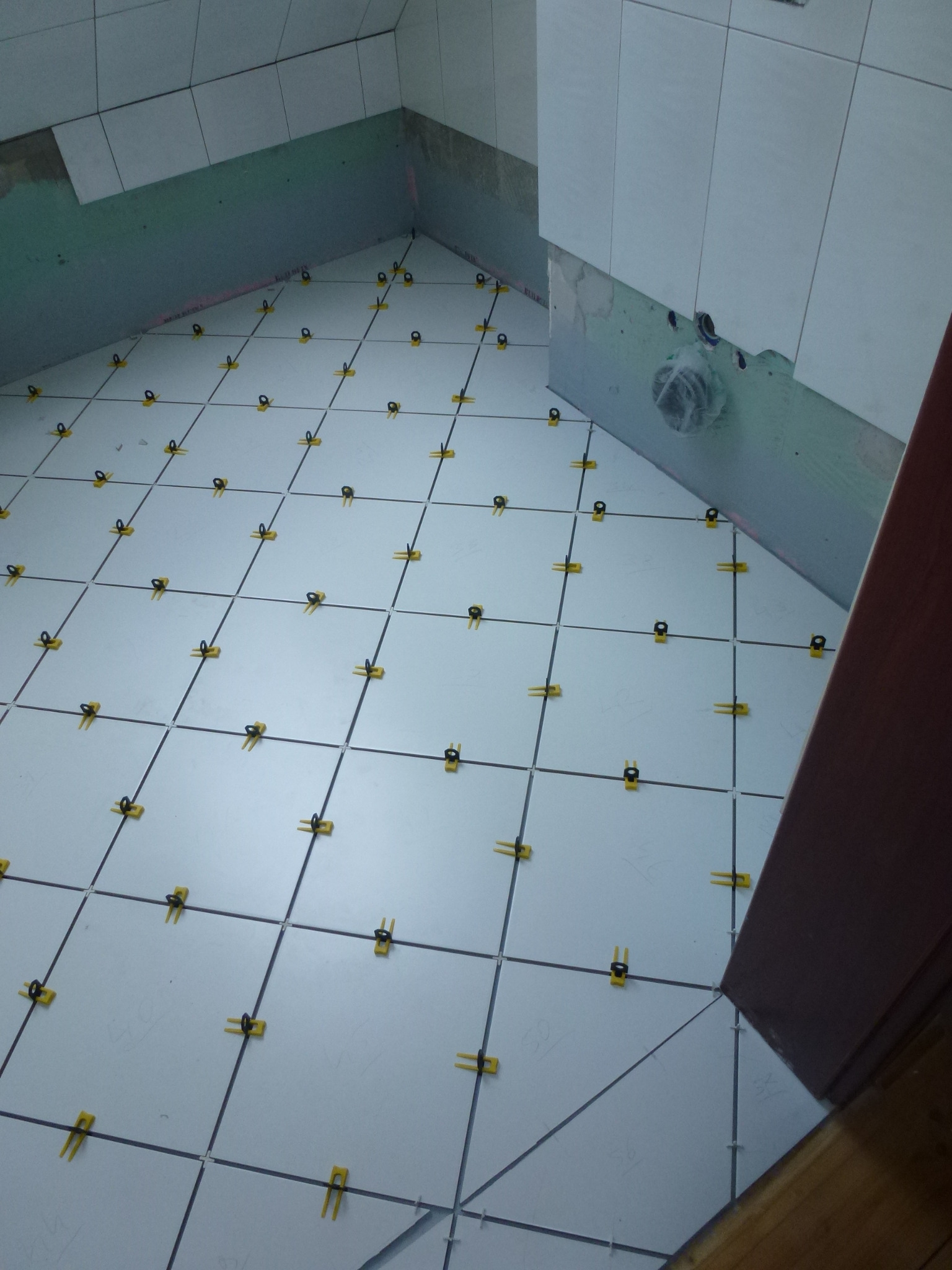 Białe płytki podłogowe w łazience, ułożone z użyciem żółtych krzyżyków dystansowych. Widoczne przygotowane ściany i fragment futryny drzwi.