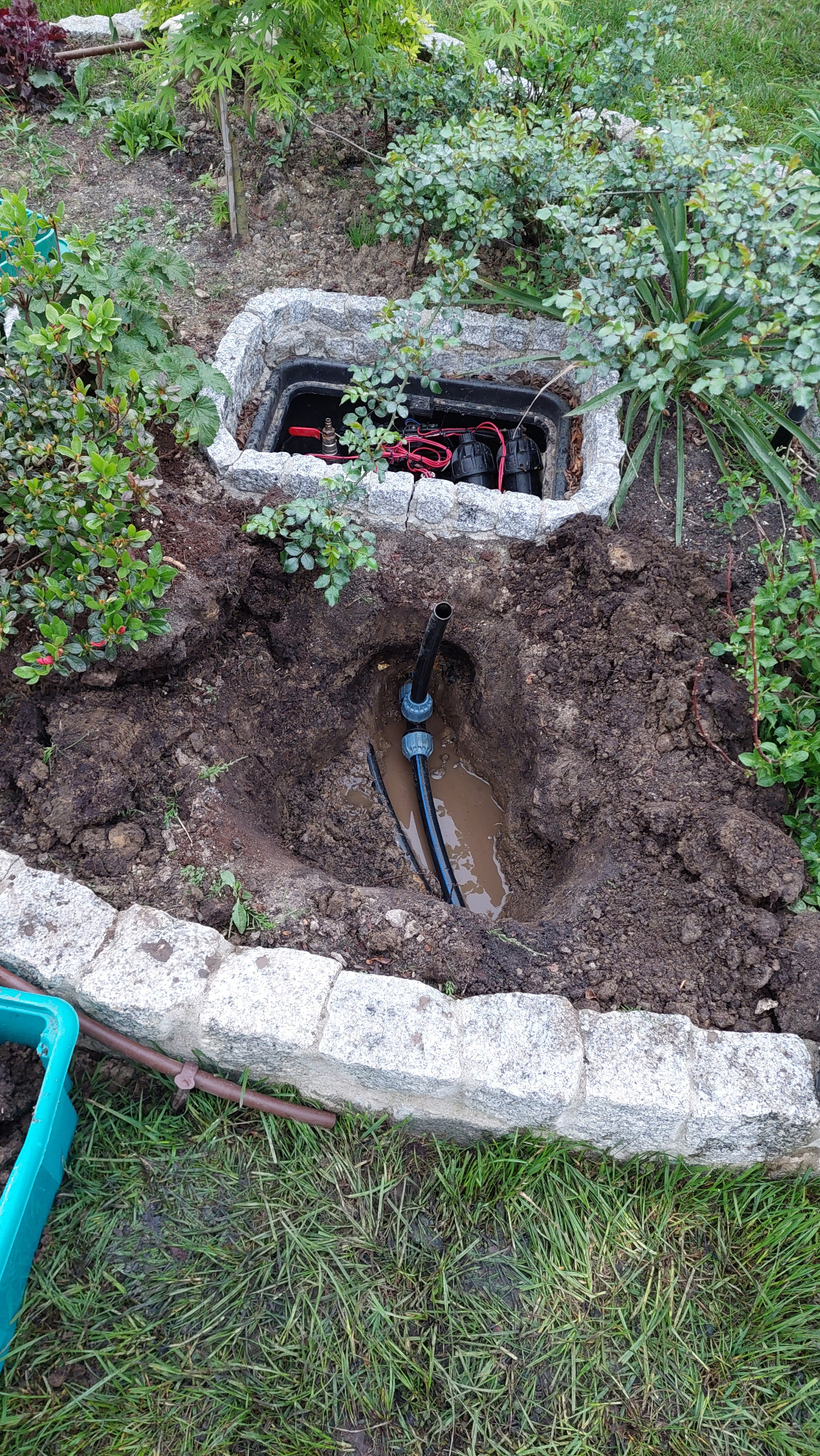 Instalacja automatycznego systemu nawadniania w Bielsko-Białej: widok na wykop z rurami i skrzynkę sterującą w kamiennej obudowie, otoczoną zielenią.