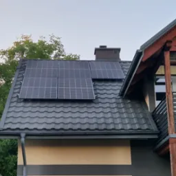 Cztery panele fotowoltaiczne na grafitowym dachu domu z kominem. Widok z dołu, z fragmentem balkonu z drewnianą konstrukcją w tle.