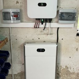 SolSpec Maciej Drozdzowski - Biały, dwuczęściowy magazyn energii Huawei, z podłączonym inwerterem i rozdzielnicami na ścianie o surowym wykończeniu. Widoczne kable i przewody elektryczne.