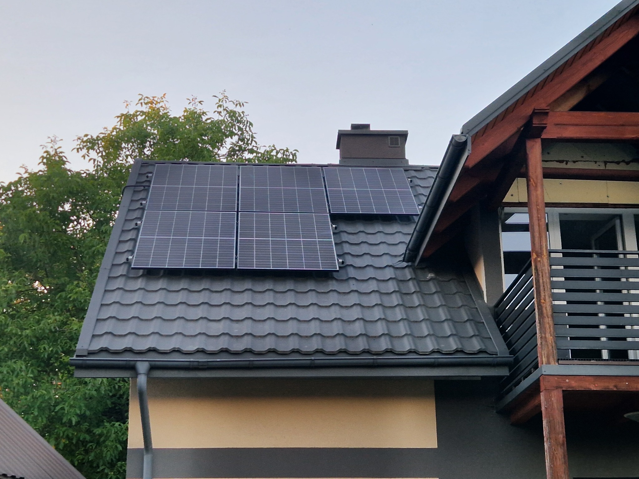 Cztery panele fotowoltaiczne na grafitowym dachu domu z kominem. Widok z dołu, z fragmentem balkonu z drewnianą konstrukcją w tle.