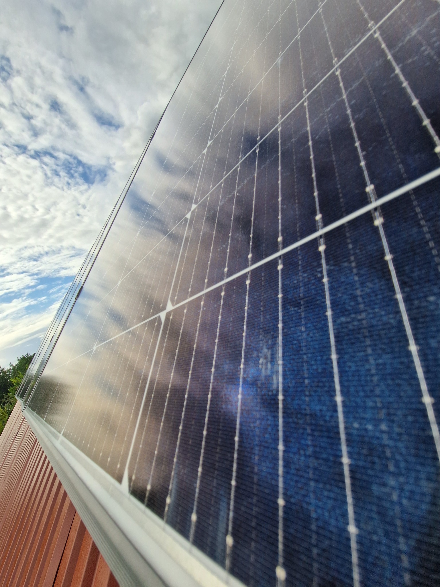 Panel słoneczny pod kątem, odbijający błękitne niebo z chmurami. Montaż na brązowej fasadzie. Widok z dołu podkreśla strukturę paneli i ich integrację z budynkiem.