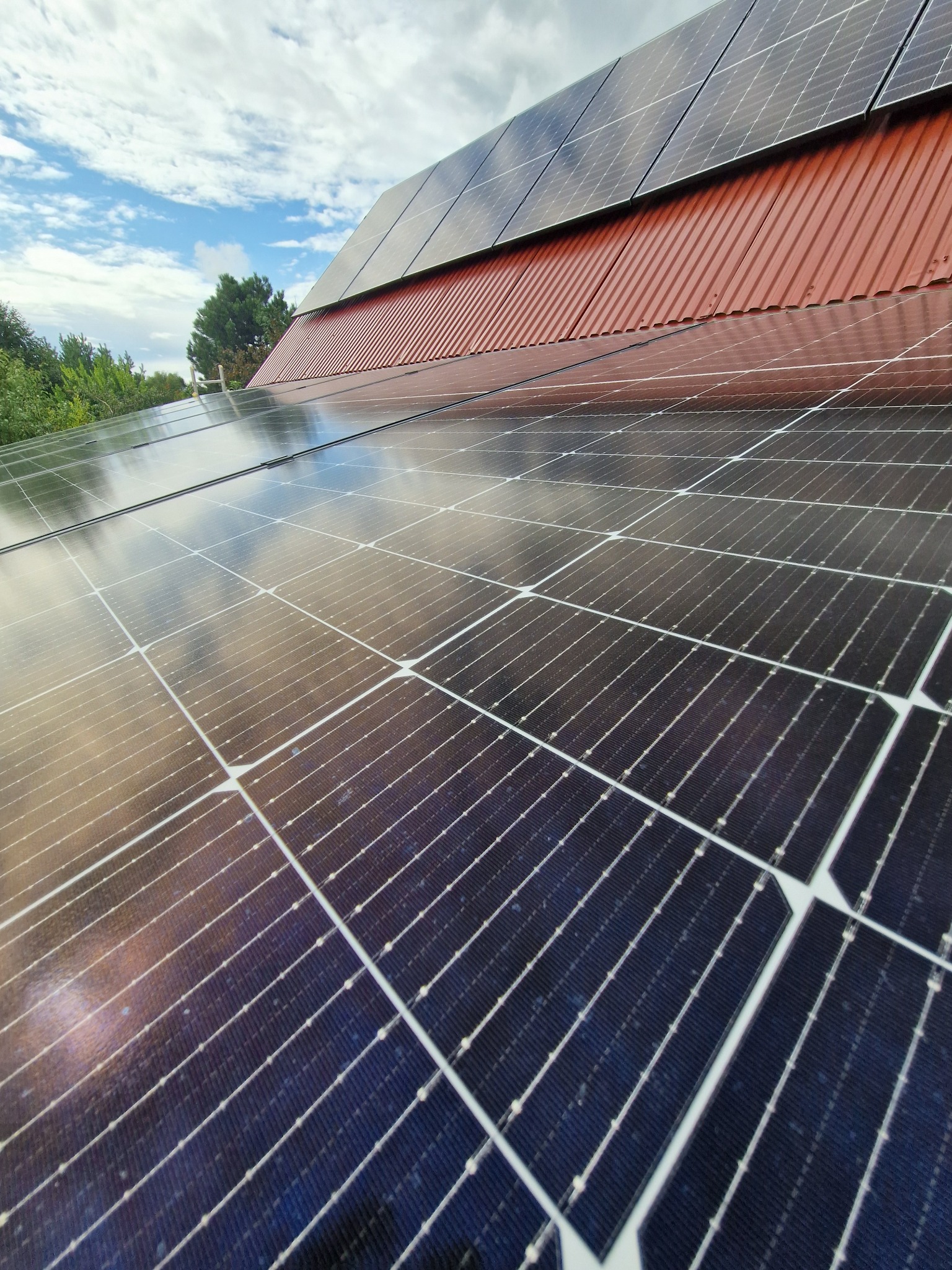 Bliższe ujęcie paneli fotowoltaicznych na dachu z czerwoną blachodachówką, odbijających błękitne niebo z chmurami. Widok pod kątem, ukazujący strukturę paneli.