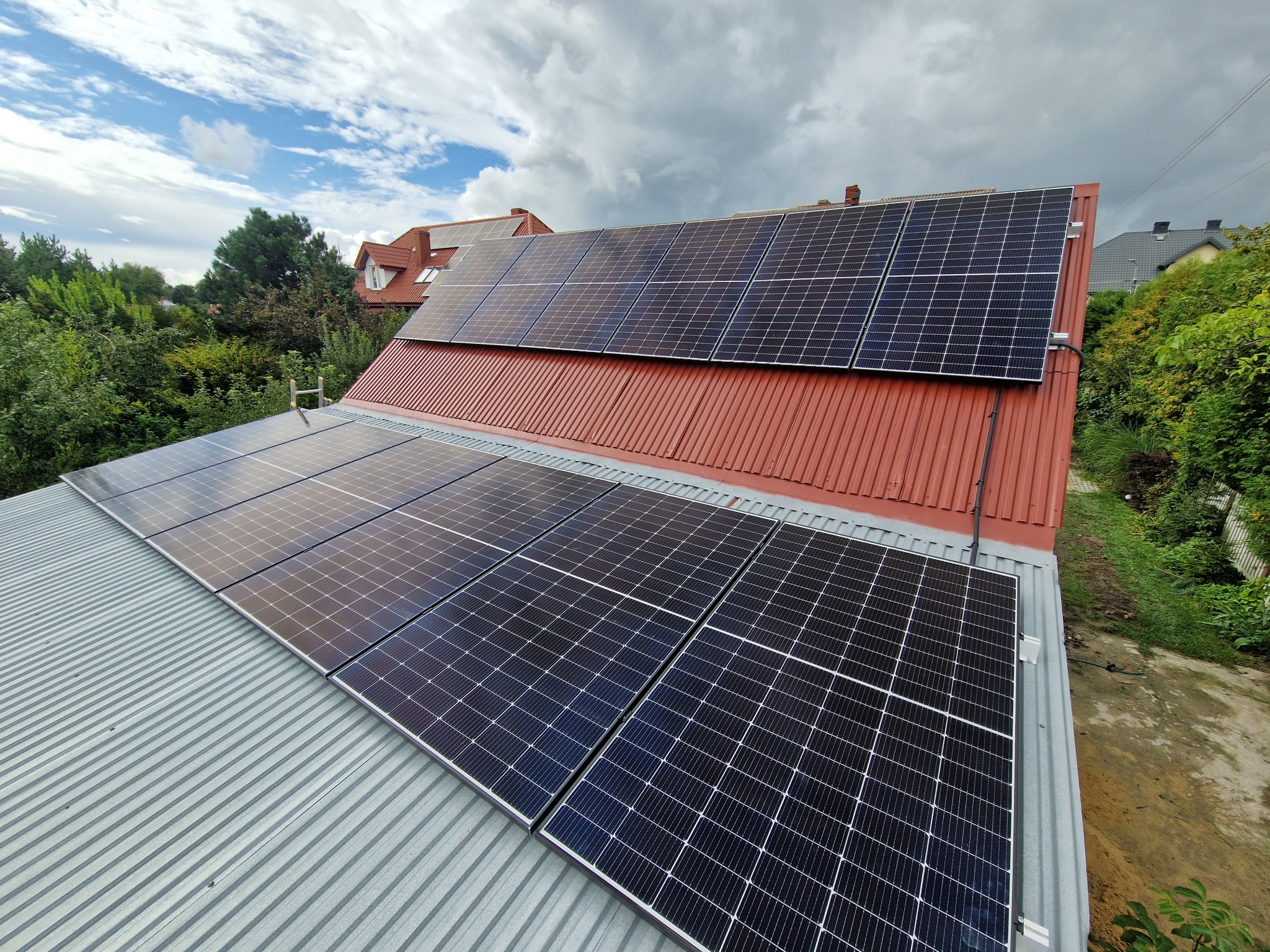Panele słoneczne na dachu z blachy trapezowej i czerwonej dachówki, widok z góry, pochmurne niebo w tle. Czarna rura biegnie wzdłuż krawędzi paneli.
