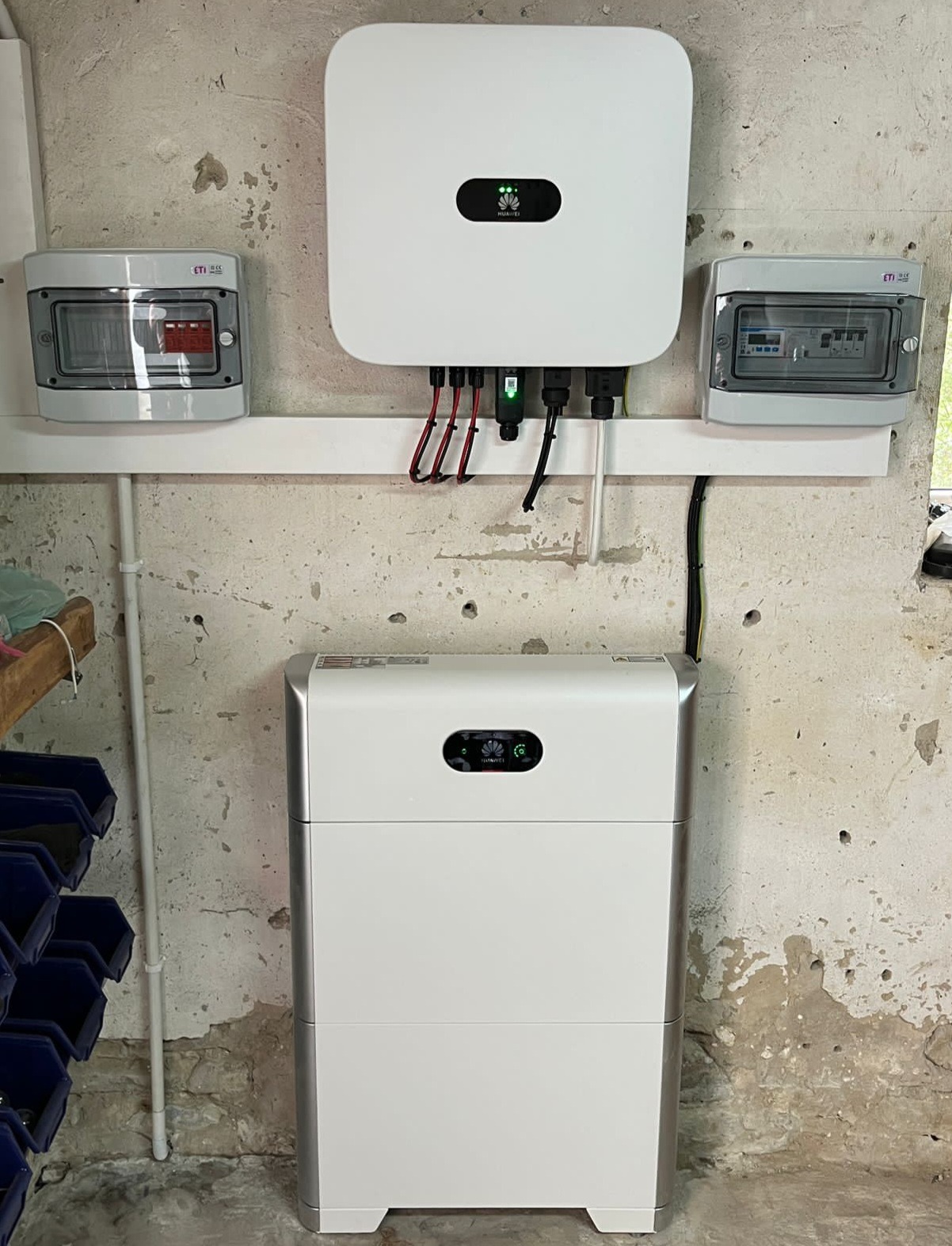 Biały, dwuczęściowy magazyn energii Huawei, z podłączonym inwerterem i rozdzielnicami na ścianie o surowym wykończeniu. Widoczne kable i przewody elektryczne.