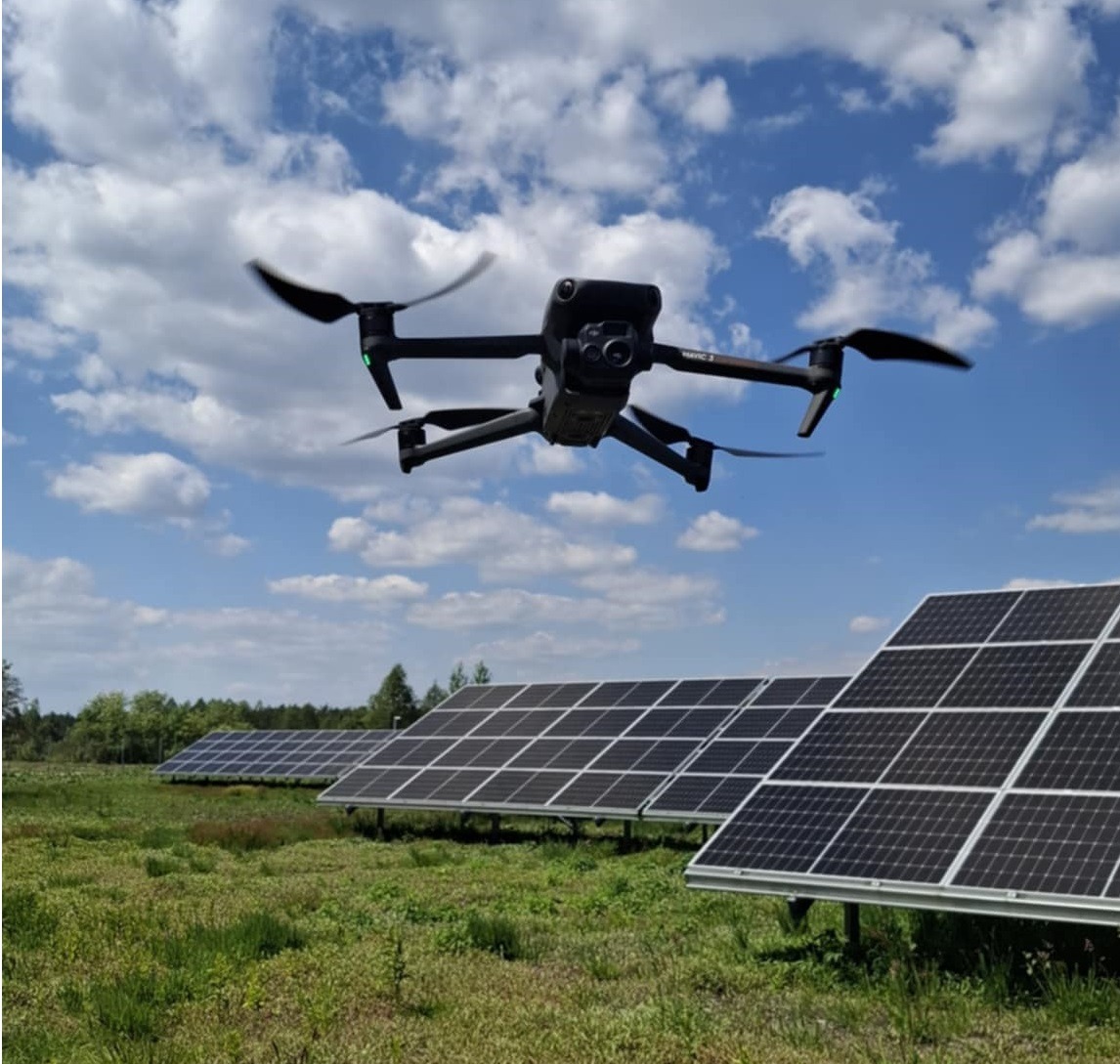 Czarny dron DJI Mavic 3 w locie nad rzędem paneli fotowoltaicznych na zielonej łące, pod błękitnym niebem z białymi chmurami. Kontrola instalacji z powietrza.