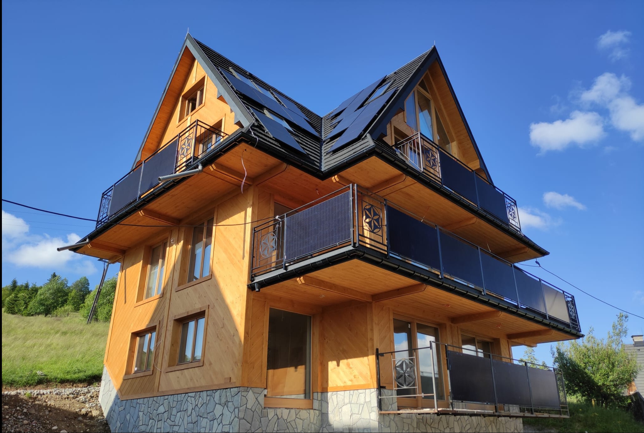 Dwupiętrowy dom z drewna z panelami fotowoltaicznymi na dachu, balkony z metalowymi balustradami. Błękitne niebo w tle.