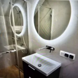 Nowoczesna łazienka: okrągłe lustro z podświetleniem LED nad umywalką z szafką, szklana kabina prysznicowa w tle. Minimalistyczny design i stonowana kolorystyka.