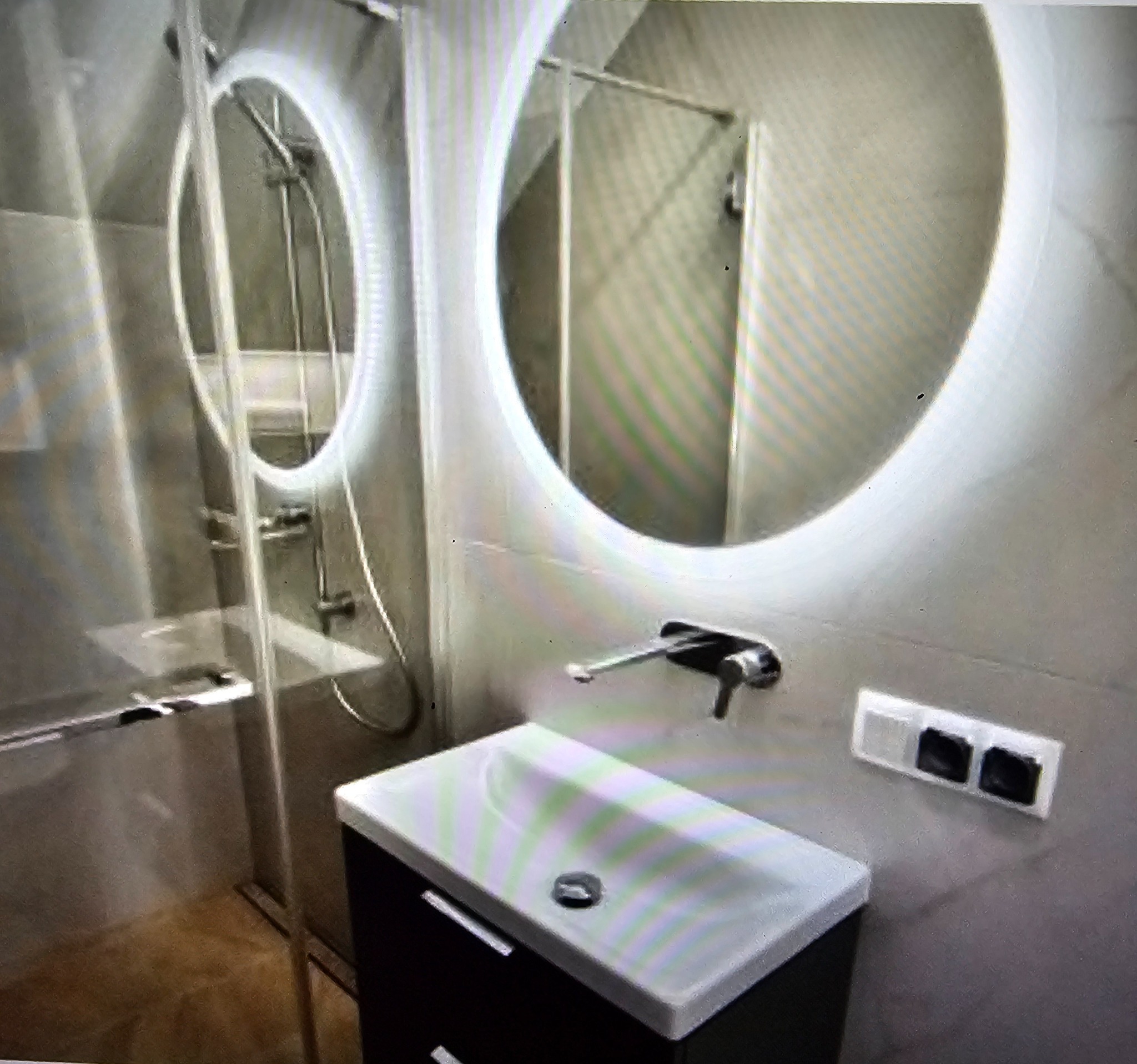 Nowoczesna łazienka: okrągłe lustro z podświetleniem LED nad umywalką z szafką, szklana kabina prysznicowa w tle. Minimalistyczny design i stonowana kolorystyka.