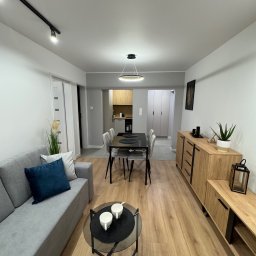 KompleksPro ALEH RADZIVONIK - Nowoczesne wnętrze mieszkania po remoncie: drewniana podłoga, szary narożnik, stół z krzesłami i drewniana komoda. Minimalistyczny design, jasne ściany, czarne akcenty.