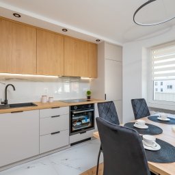 KompleksPro ALEH RADZIVONIK - Jasna kuchnia z drewnianymi szafkami, czarną armaturą i marmurowym backsplashiem. Stół z nakryciem na cztery osoby, krzesła z pikowaniem. Minimalistyczna lampa.