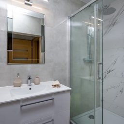 KompleksPro ALEH RADZIVONIK - Jasna łazienka z kabiną prysznicową typu walk-in, białą umywalką z szafką i lustrem. Ściany wyłożone płytkami w odcieniach szarości i bieli imitującymi marmur.