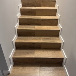 KompleksPro ALEH RADZIVONIK - Schody wyłożone płytkami imitującymi drewno, z białymi listwami przypodłogowymi, widok z dołu, symetryczny układ, jasne oświetlenie, minimalistyczny design.