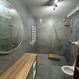 KompleksPro ALEH RADZIVONIK - Szara łazienka z marmurowymi płytkami, okrągłym lustrem i drewnianą półką. Widoczny prysznic typu walk-in, grzejnik drabinkowy i oświetlenie sufitowe.