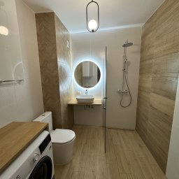KompleksPro ALEH RADZIVONIK - Nowoczesna łazienka z drewnopodobnymi płytkami, okrągłym lustrem z podświetleniem LED, pralką i kabiną prysznicową typu walk-in. Minimalistyczny design i ciepła kolorystyka.