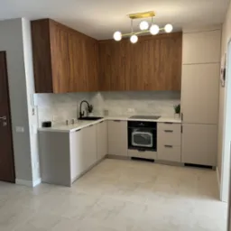 Nowoczesna kuchnia w beżach i brązach z marmurowym blatem, oświetlona złotą lampą sufitową. Uwagę zwraca czarna armatura i minimalistyczne dodatki.