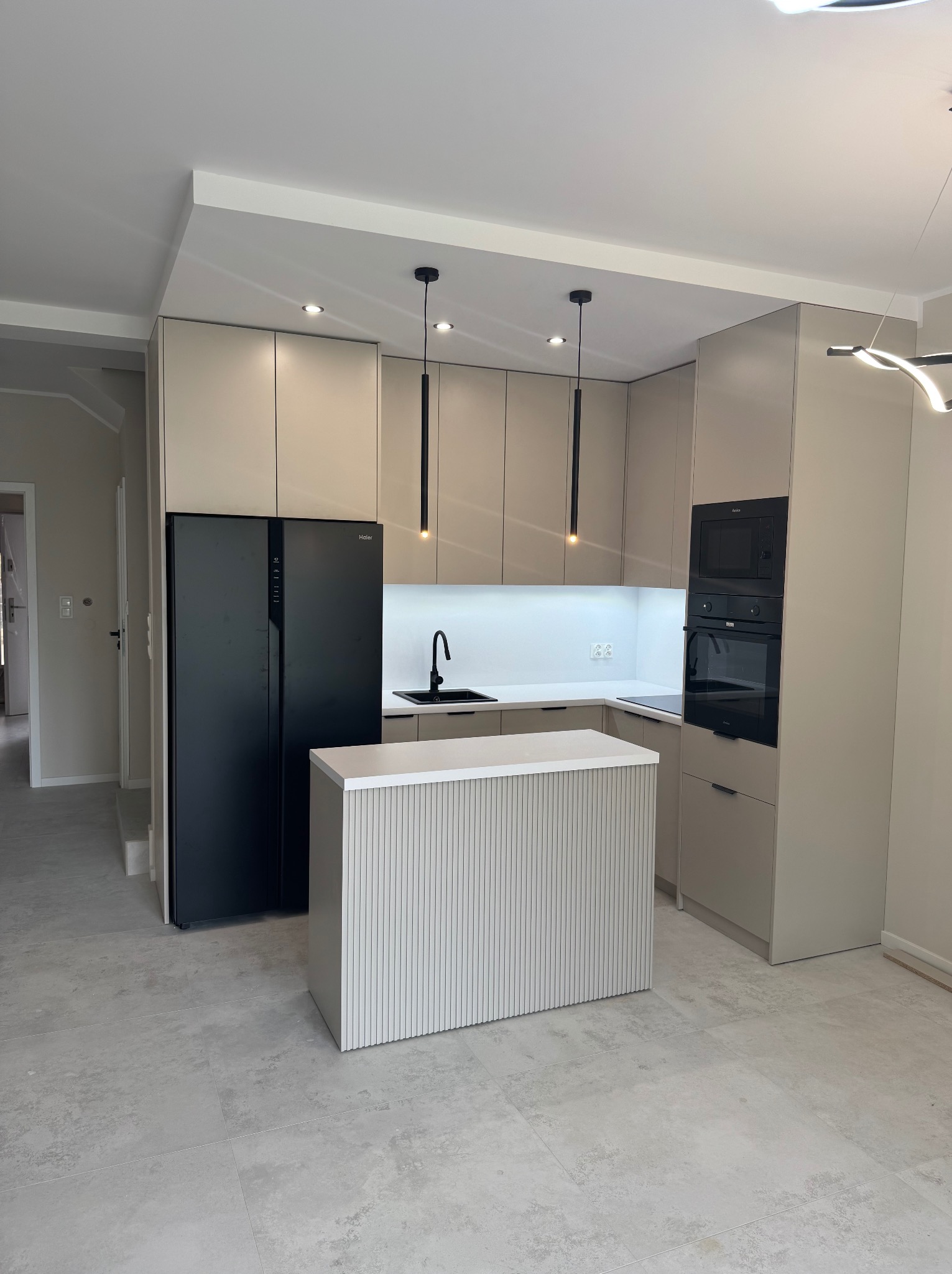 Nowoczesna kuchnia w odcieniach beżu z czarnymi akcentami: lodówka side-by-side, wyspa z ryflowanym frontem, oświetlenie punktowe i wiszące. Minimalistyczny design.