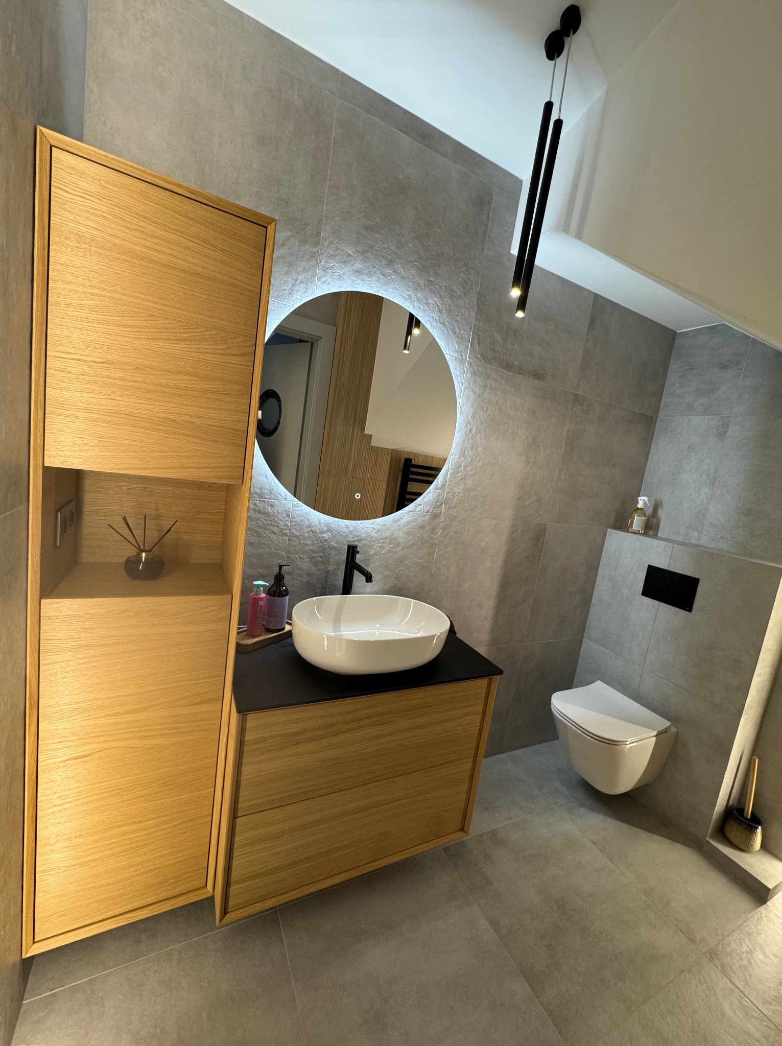 Nowoczesna łazienka z szarą glazurą, drewnianą szafką pod umywalką, okrągłym lustrem z podświetleniem LED i wiszącą toaletą. Minimalistyczny design.