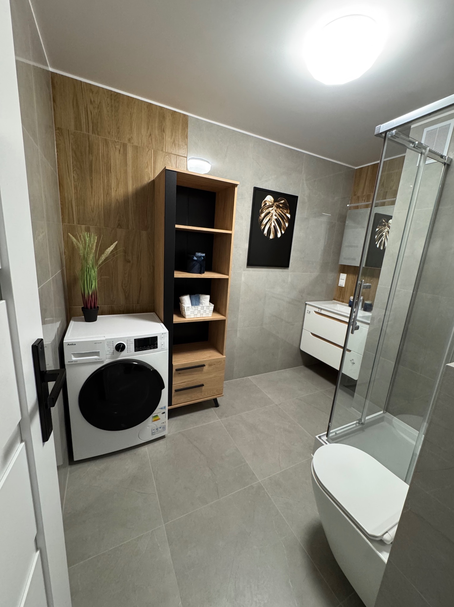 Nowoczesna łazienka w szarościach i drewnie: kabina prysznicowa, umywalka z szafką, pralka i regał. Na ścianie dekoracja z motywem liścia.