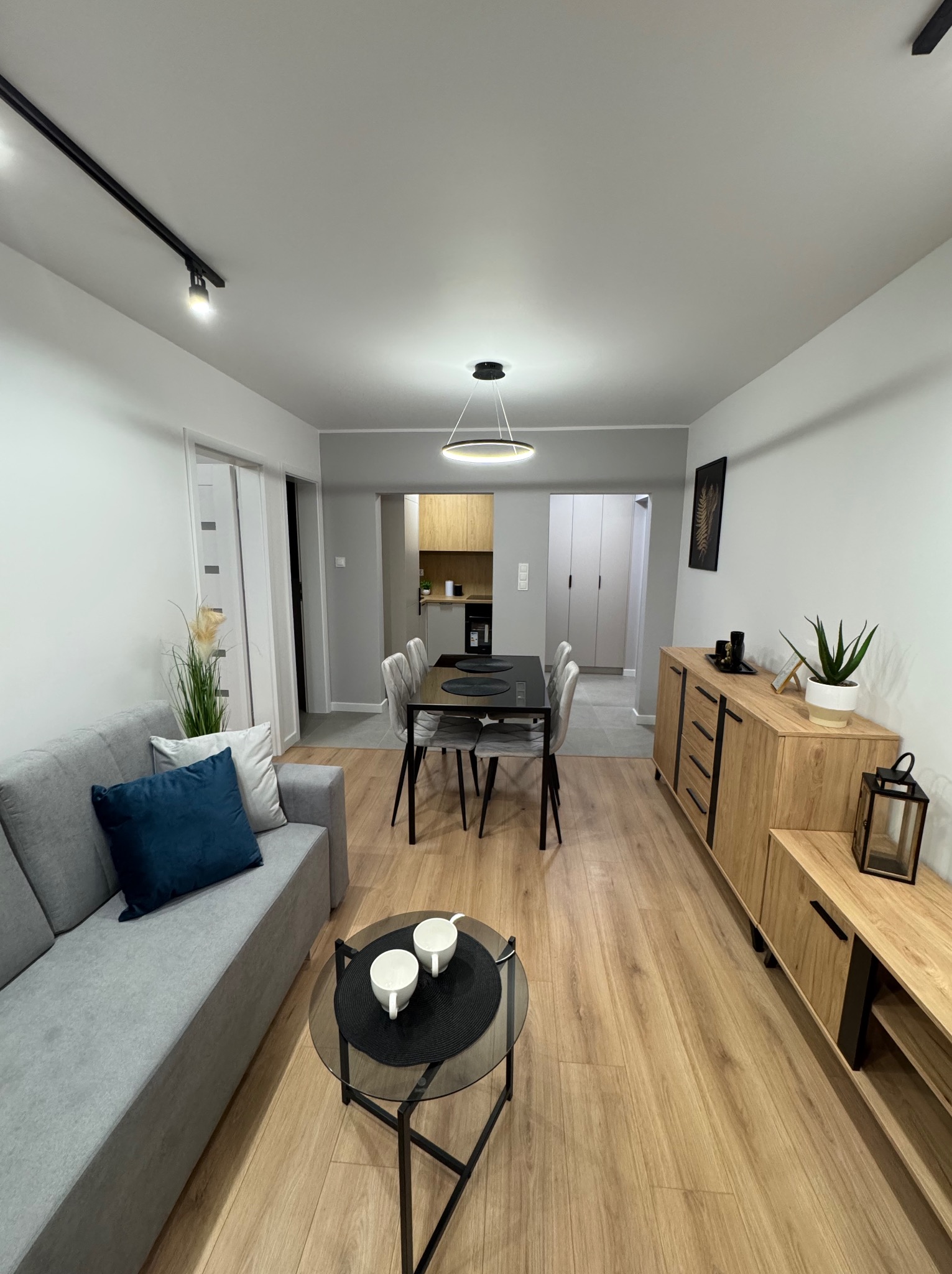 Nowoczesne wnętrze mieszkania po remoncie: drewniana podłoga, szary narożnik, stół z krzesłami i drewniana komoda. Minimalistyczny design, jasne ściany, czarne akcenty.