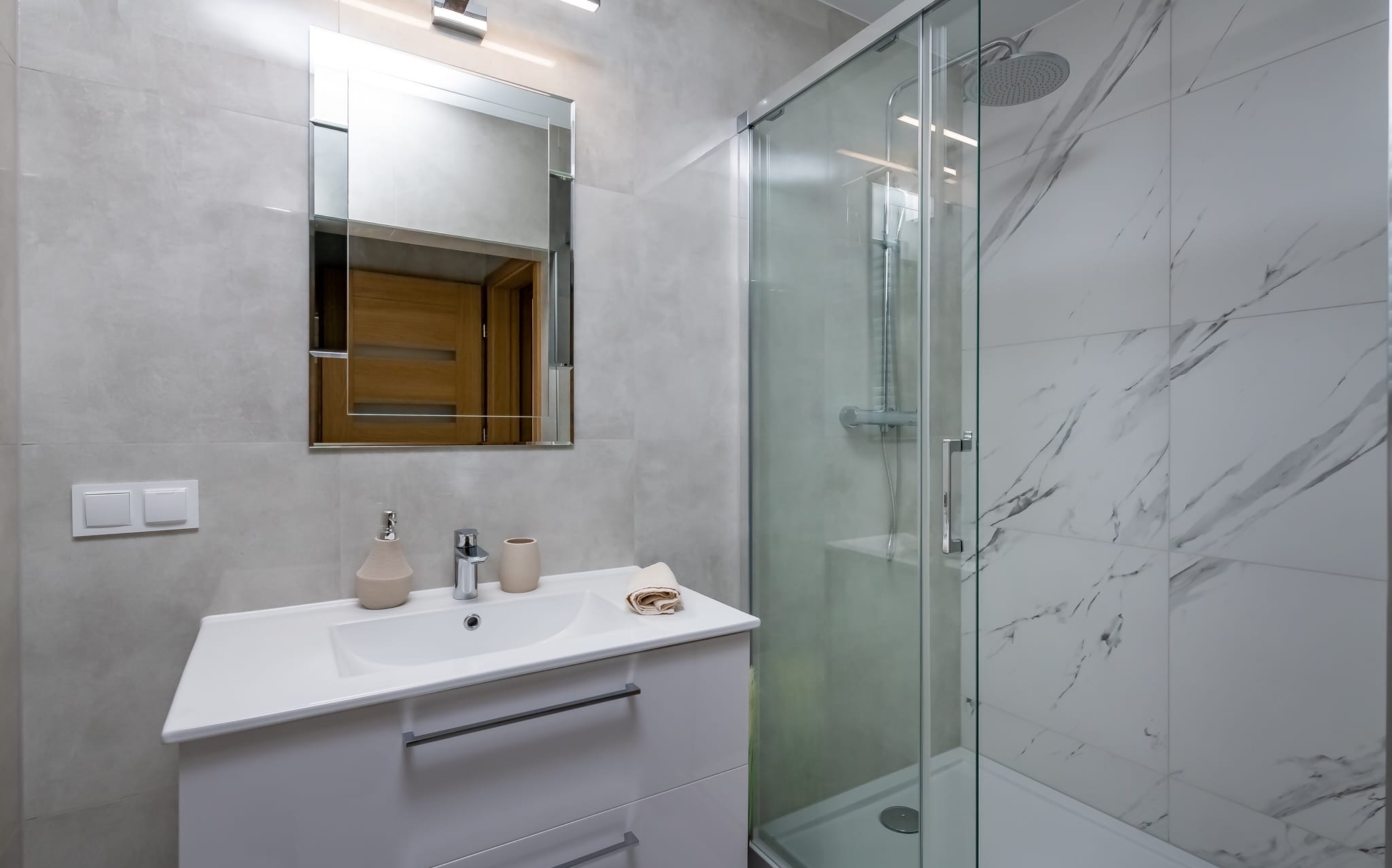 Jasna łazienka z kabiną prysznicową typu walk-in, białą umywalką z szafką i lustrem. Ściany wyłożone płytkami w odcieniach szarości i bieli imitującymi marmur.