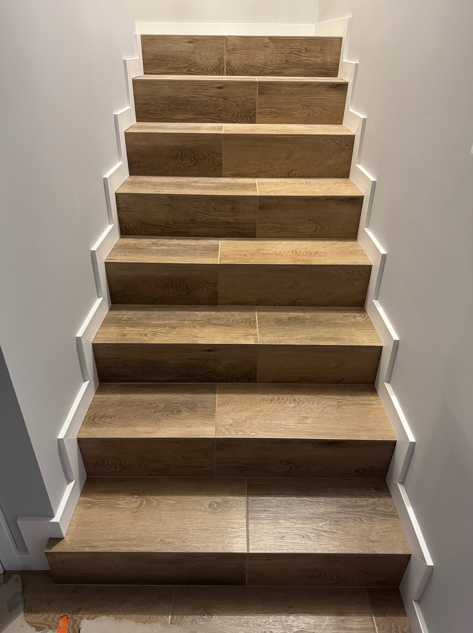 Schody wyłożone płytkami imitującymi drewno, z białymi listwami przypodłogowymi, widok z dołu, symetryczny układ, jasne oświetlenie, minimalistyczny design.