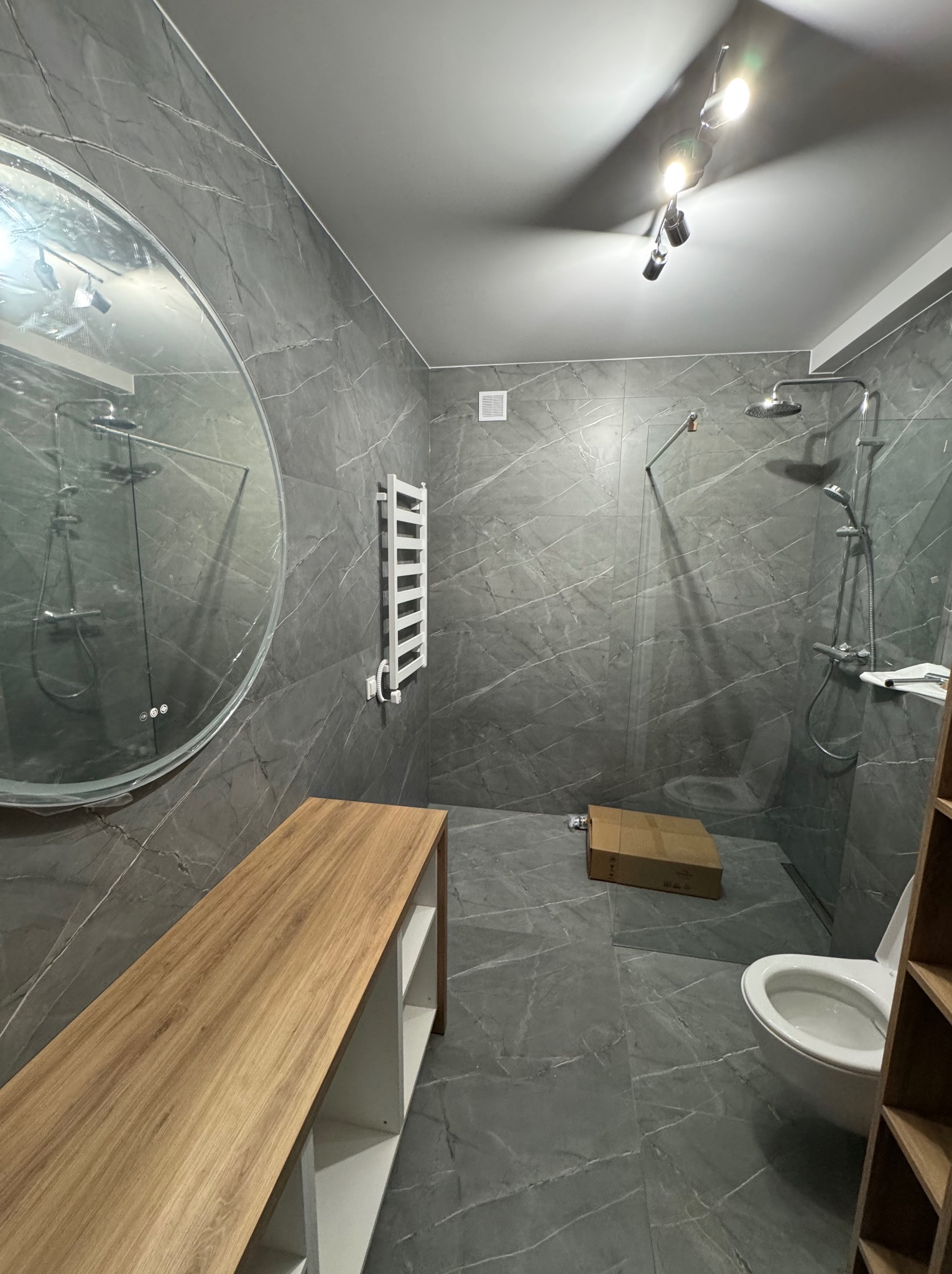 Szara łazienka z marmurowymi płytkami, okrągłym lustrem i drewnianą półką. Widoczny prysznic typu walk-in, grzejnik drabinkowy i oświetlenie sufitowe.