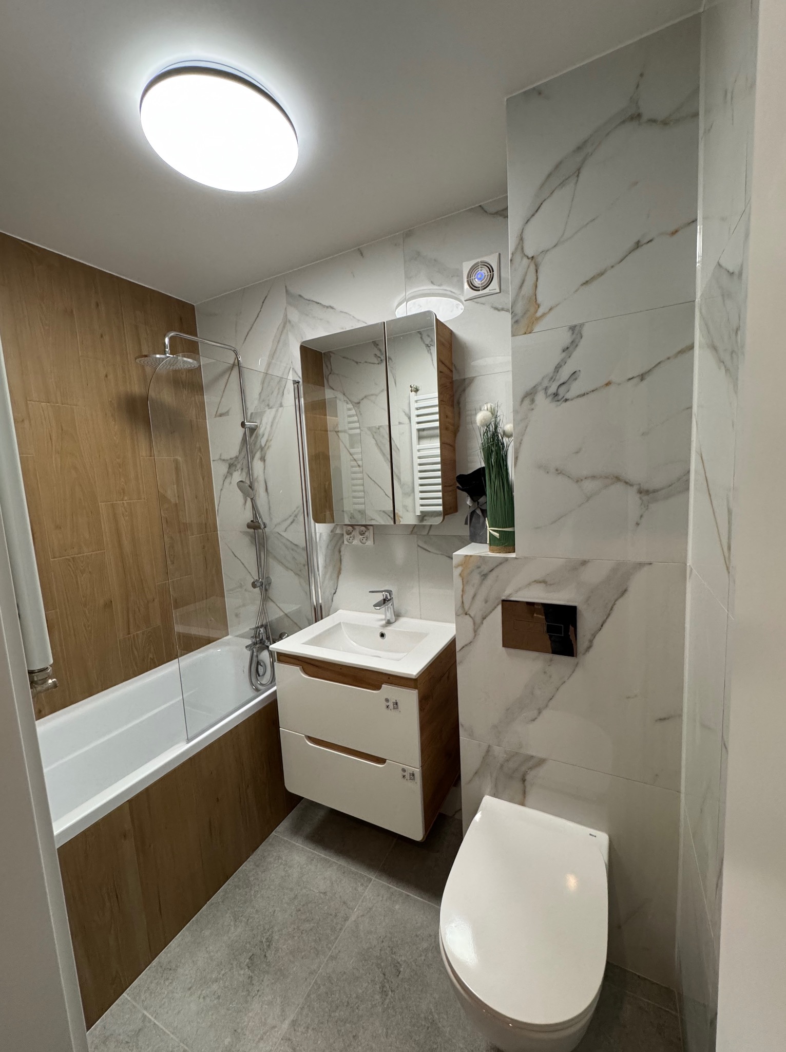 Nowoczesna łazienka po remoncie: marmurowe płytki, drewnopodobne akcenty, biała armatura i szara podłoga. Lustro z oświetleniem nad umywalką.