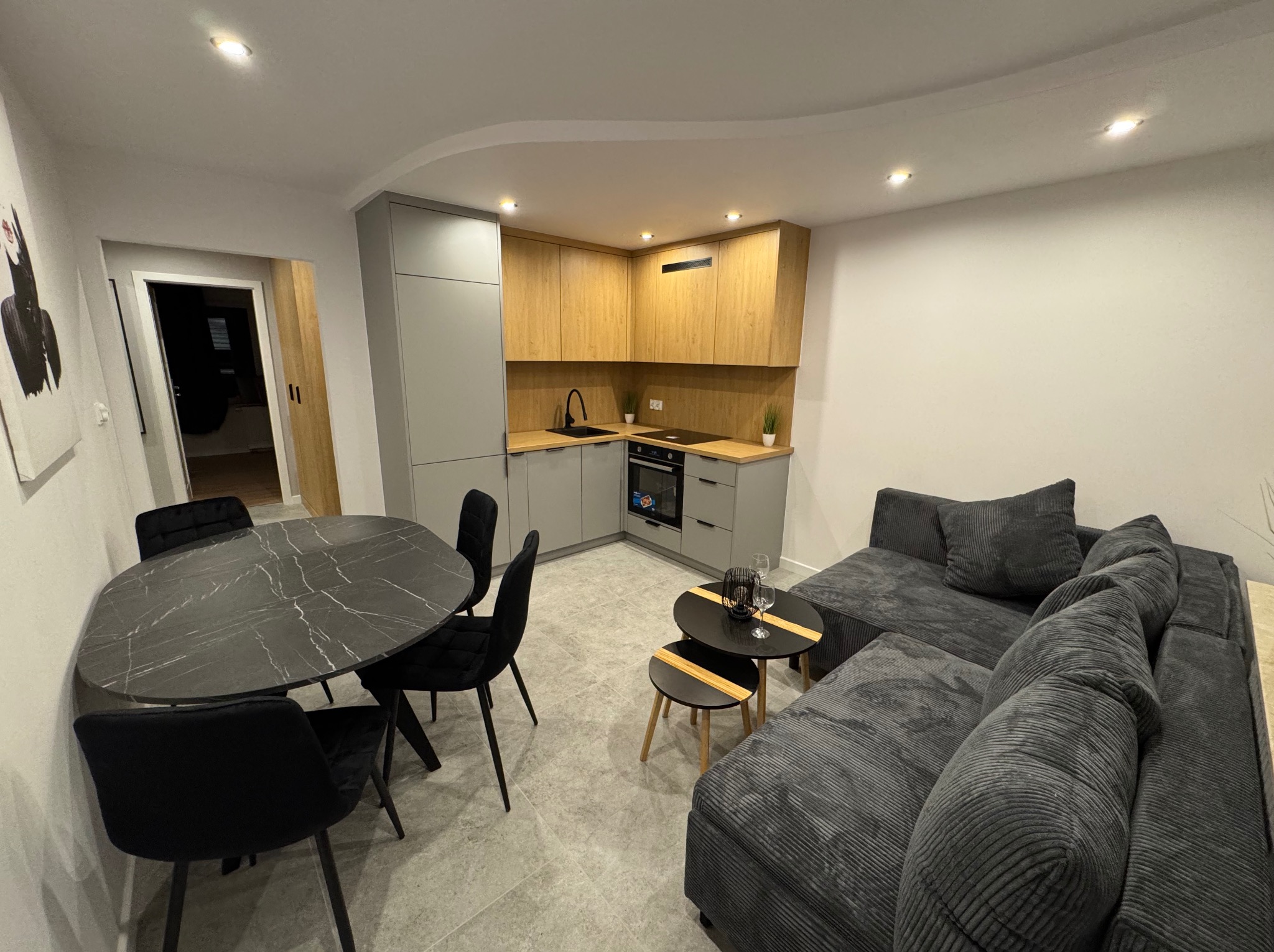 Nowoczesne wnętrze: szara kuchnia z drewnianymi akcentami, czarny stół z krzesłami i szara sofa w przestronnym salonie. Widok na aranżację po remoncie.