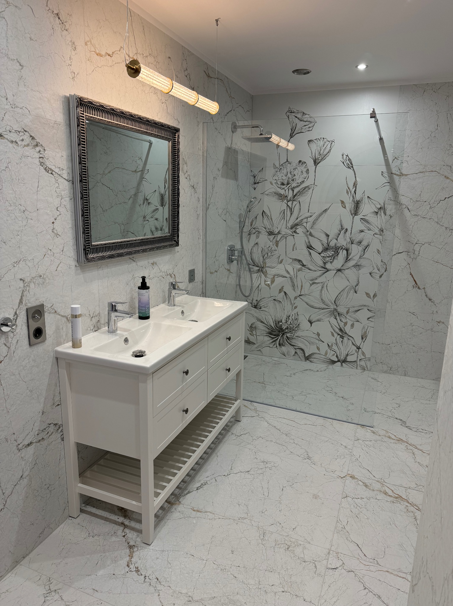 Elegancka łazienka z marmurowymi płytkami, białą szafką pod umywalką i kabiną prysznicową z motywem kwiatowym. Lustro w ozdobnej ramie i nowoczesne oświetlenie.